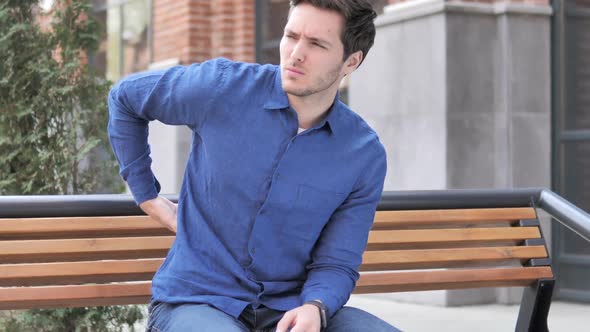 Young Man with Back Pain Sitting Outdoor on Bench alt