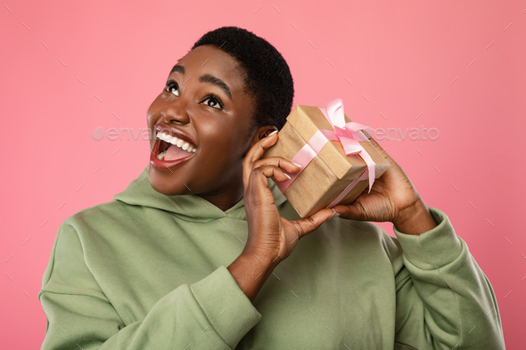 Happy Black Lady Shaking Gift Box Near Ear, Pink Background