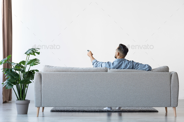 Man Sitting On Couch Opening Air Conditioner With Remote Controller, Rear View
