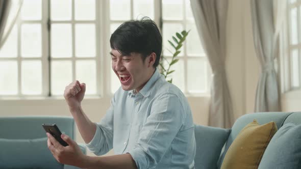 Asian Male Using Smartphone And Celebrating In Living Room alt