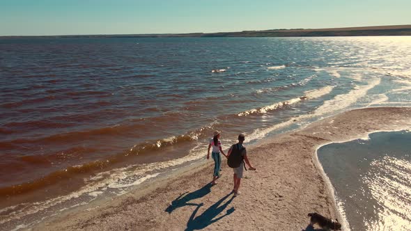 Aerial View From a Drone Flying Around of Happy Couple and Dog Walking By Sand Beach Between Water alt