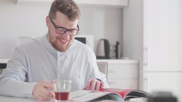 A Young Stylish Man with Glasses in the Morning at Breakfast Reads a Magazine and Drinks Tea