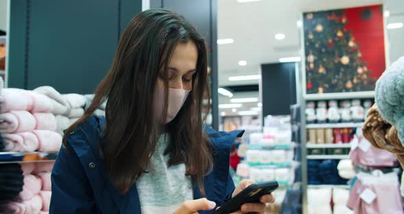 Young Beautiful Woman Using Phone During Shopping in Store and Wearing Face Mask alt