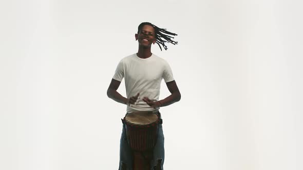 Black Man with Dreadlocks Cheerfully Plays an African Folk Musical Drum Instrument on White alt