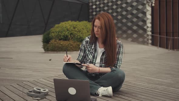 Caucasian Redhaired Girl Sits on a Wooden Floor Makes Plans Using a Notebook and a Computer