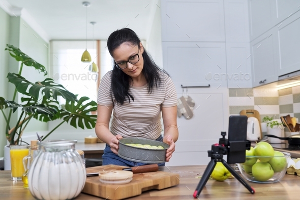 Woman preparing apple pie at home in the kitchen, with a smartphone using a video call
