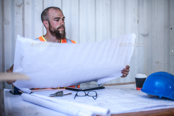 Construction worker sitting at desk looking at blueprint in portable cabin