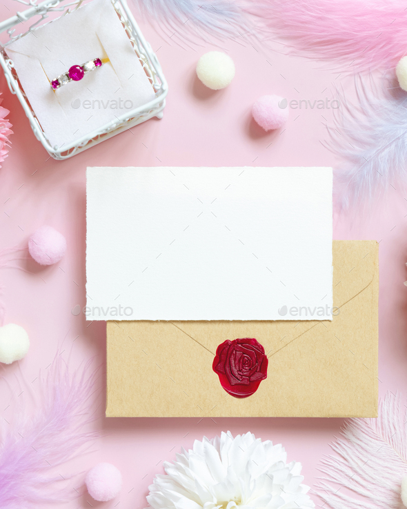 Card and envelope near pastel flowers, pom-poms, feathers and ring in a gift box on pink