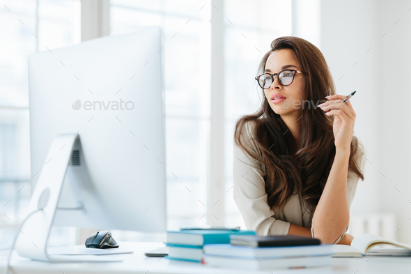 Dark haired lady focused into big monitor, sits at desktop, holds pen and writes notes