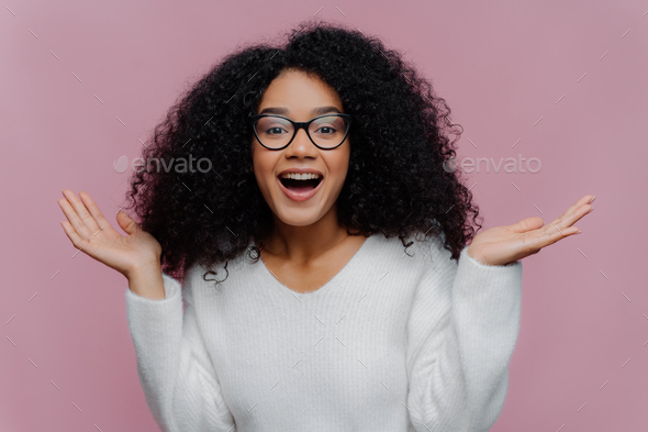 Joyful Curly Lady Raises Arms And Spreads Palms Wears Optical Glasses And White Sweater Stock