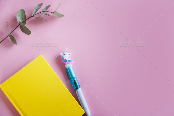 Yellow notebook for notes, funny unicorn pen and green eucalyptus leaves on pink pastel background