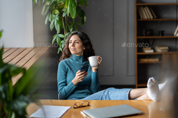 Woman social media manager work from home sit at closed laptop, feet on desk drink morning coffee