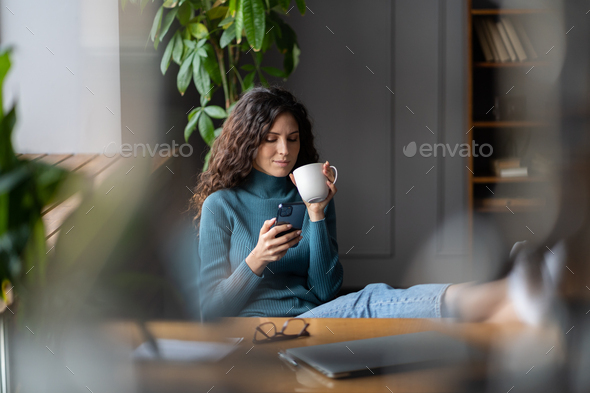 Happy woman procrastinating at workplace drink coffee read online news on smartphone at office desk