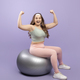 Happy young european plus size woman in sports uniform shows biceps on arms, sits on fitness ball - PhotoDune Item for Sale