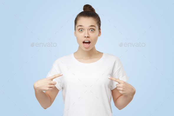 Good-looking young woman points to herself with eyes and mouth round with surprise