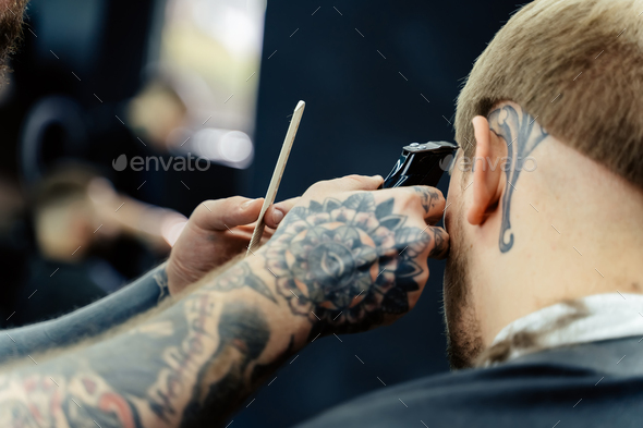 Male haircut with electric razor. Tattooed Barber makes haircut for client  at the barber shop by Stock Photo by licsiren