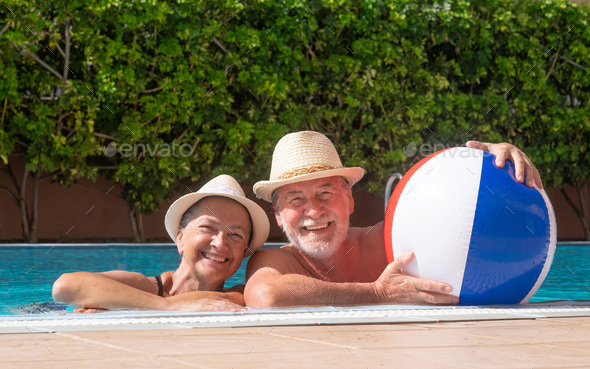 Happy adult senior caucasian couple laughing looking at camera floating in outdoor swimming pool