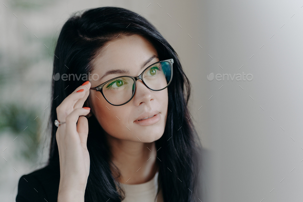 Brunette serious lady keeps hand on rim of spectacles, concentrated at computer screen