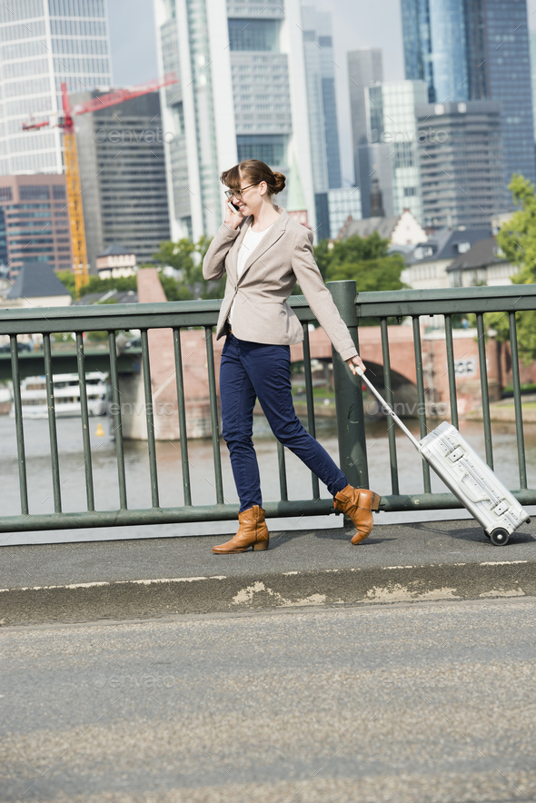 Germany, Hesse, Frankfurt, telephoning businesswoman walking with rolling suitcase over a bridge