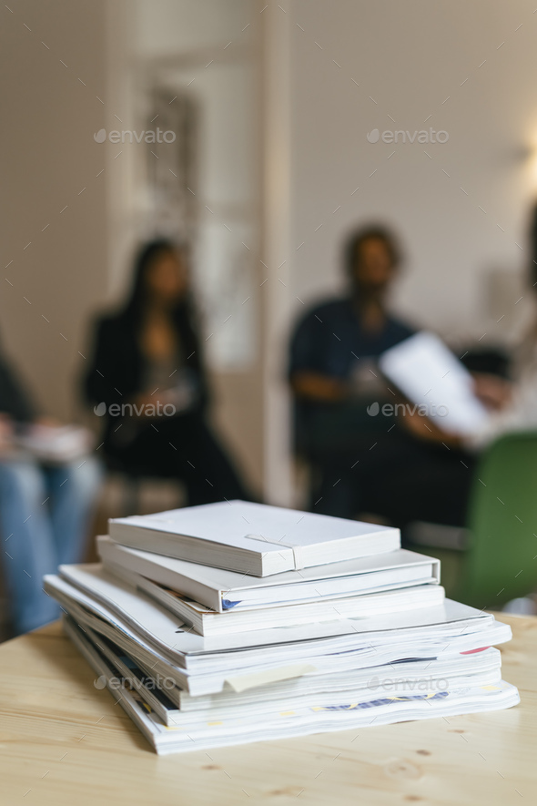 Pile of magazines and books on shelf, business peole talking in background