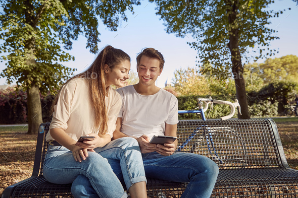 Young couple in park sitting on a bench with cell phone and tablet