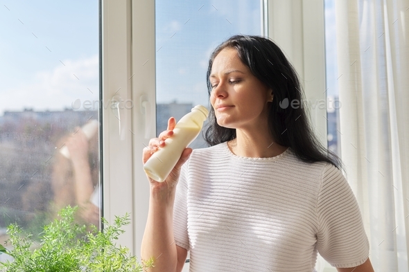 Woman drinking milk drink from bottle standing near window, milk yogurt dairy healthy drinks