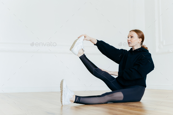 Healthy female works out in spacious hall, raises leg and touches toe, wears black hoody
