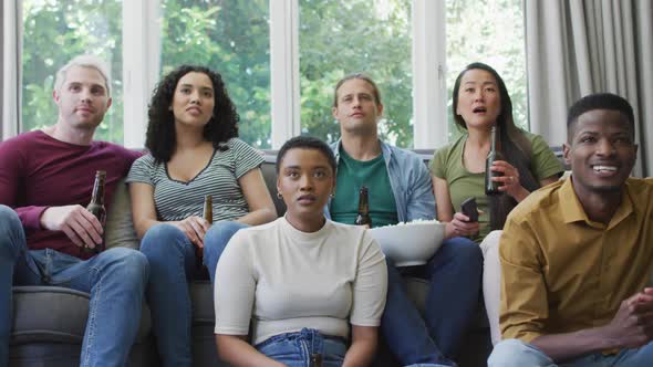 Diverse group of happy male and female friends watching sport drinking beer, cheering in living room alt