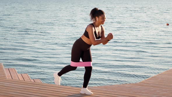 Young Sporty Black Woman Exercising with Elastic Band Listening to Music in Earbuds Working Out on alt