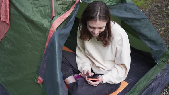 A Young Tourist Sits in a Tent in Nature and Looks at the Phone