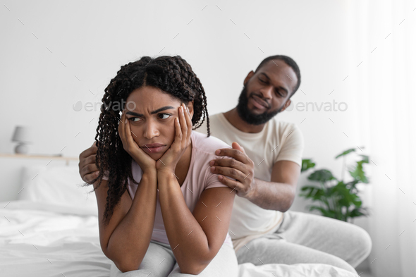 Millennial african american husband calm his wife, sit on comfort bed in bedroom interior