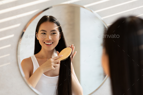 Cheerful asian lady brushing hair, standing in front of mirror