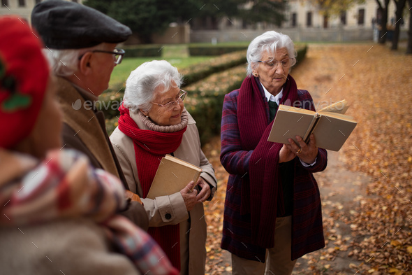 Group of happy senior friends with book on walk outdoors in park in autumn, reading and talking - Stock Photo - Images