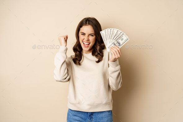 Enthusiastic modern woman winning money, got cash, celebrating and shouting of joy, standing against