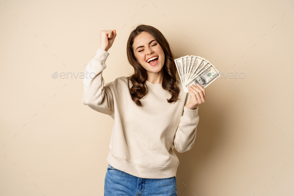 Enthusiastic modern woman winning money, got cash, celebrating and shouting of joy, standing against