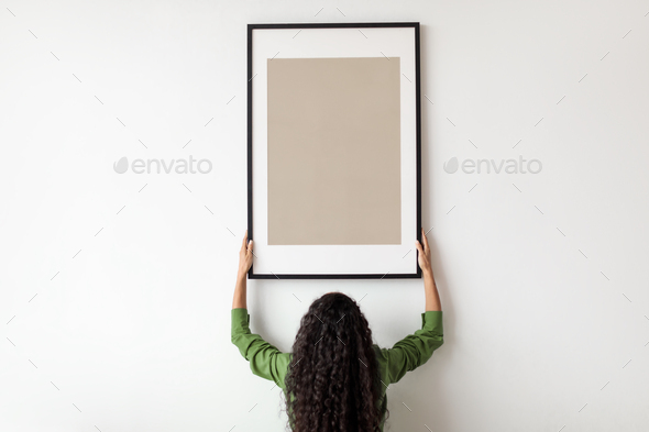 Unrecognizable Female Hanging Blank Poster In Frame On Wall Indoor