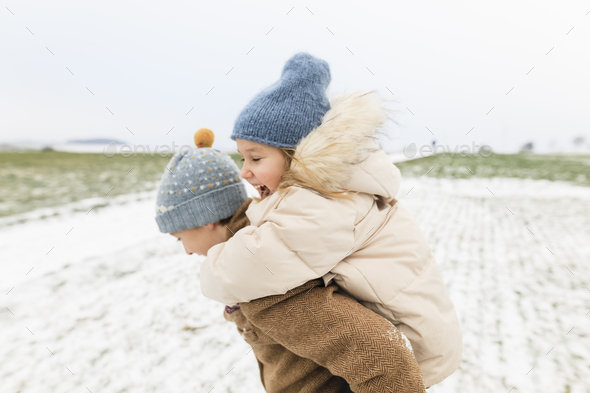 Boy carrying happy sister piggyback in winter landscape