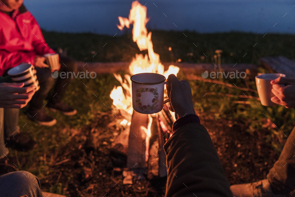 Hand of person holding tea cup, group of people sitting at a camp fire