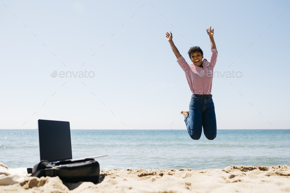 Woman jumping on the beach, laptop on bag
