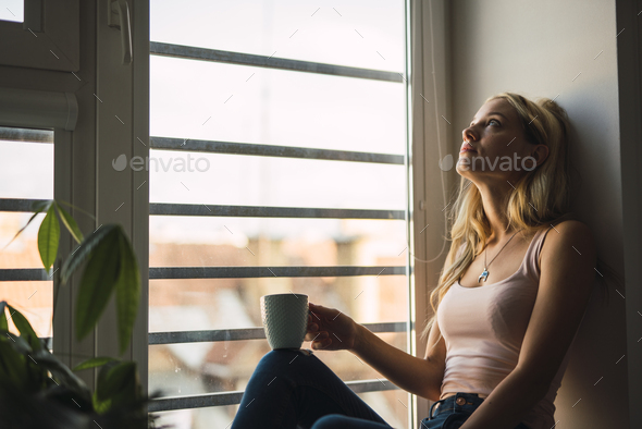 Blond Young Woman Holding Coffee Mug Sitting In Windowsill Stock Photo