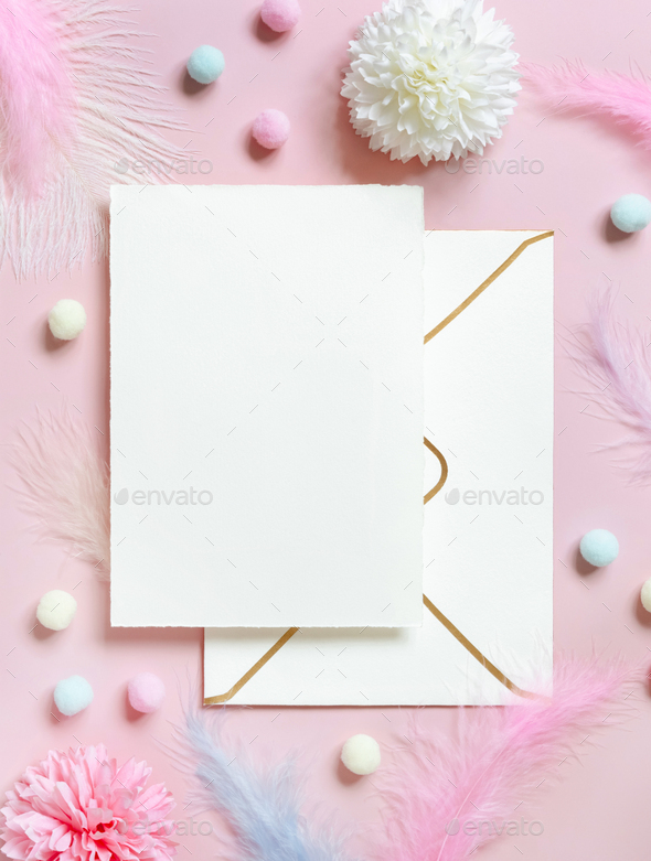 Blank card and envelope near pastel flowers, pom-poms and feathers near ring in a gift box on pink