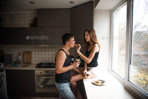 Young millennial couple is sitting on the windowsill near window, drinking latte coffee with cookies - Stock Photo - Images
