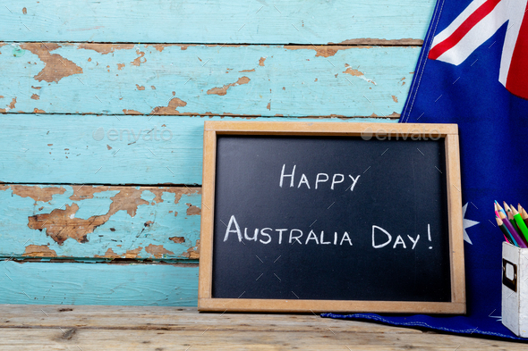 Happy australia day text on writing slate by national flag against wooden wall with copy space