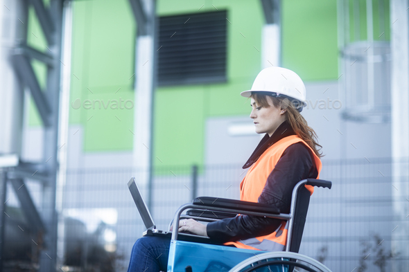 Young technician with safety helmet and vest in wheelchair working on laptop outdoors