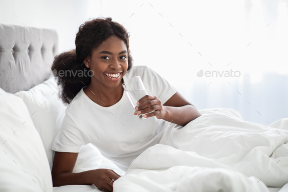 Smiling pretty black lady drinking water in bed, copy space