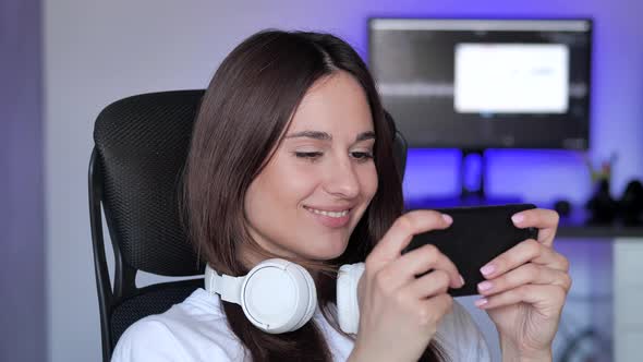 Cute Gamer Woman Sit on a Gaming Chair Smiling and Playing Mobile Online Game on a Smartphone alt