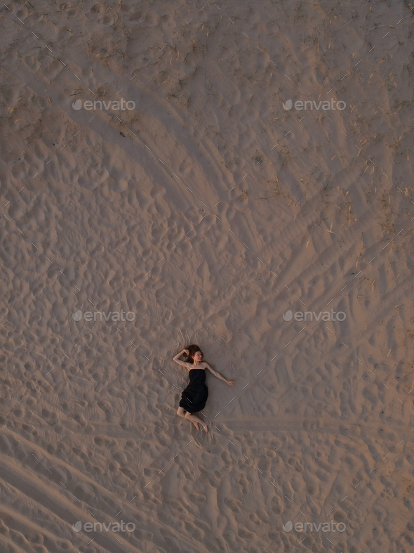 Young woman in a black dress lying on the back on the sand. Top view from the drone.