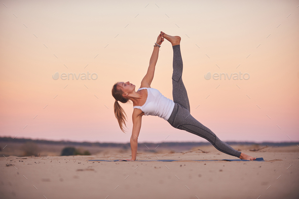 Beautiful young woman practices yoga advanced side plank pose vasisthasana in the desert at sunset