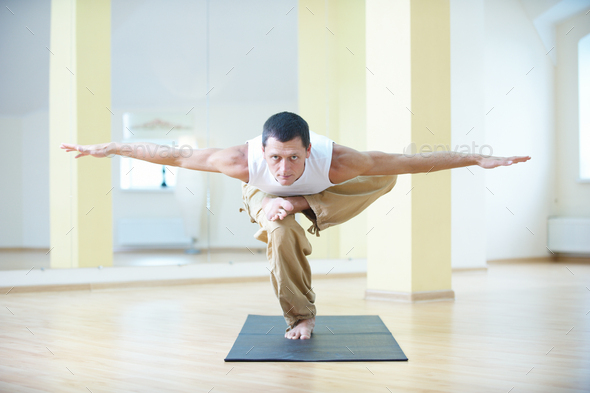 A young strong man stands in the asana on one leg with the arms apart in the yoga studio