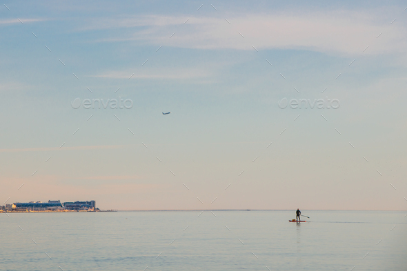 A lone man with a bag moving across the sea on a surfboard with an oar. Minimalism